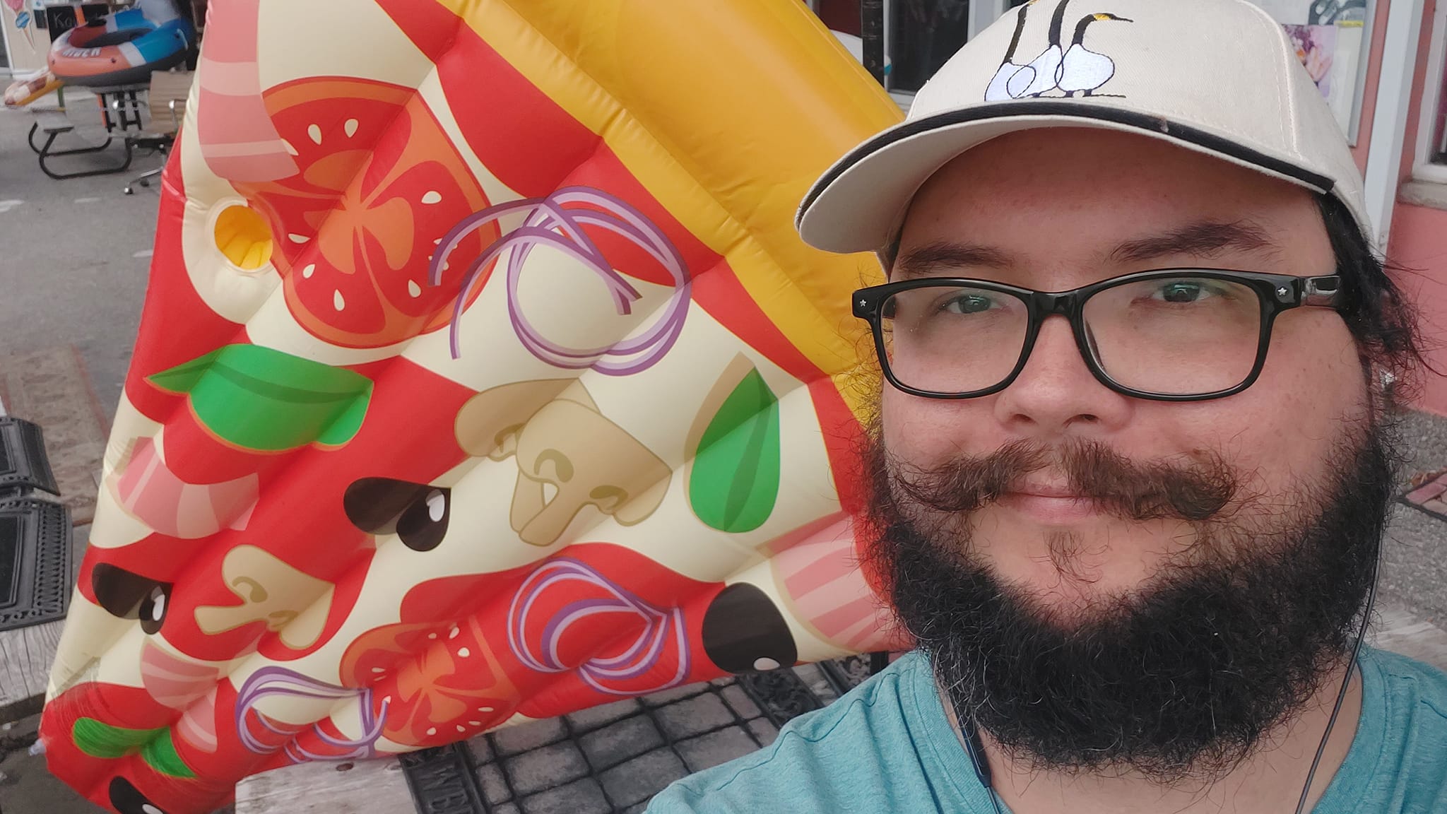 A man with short hair and bushy facial hair stands in front of a giant inflatable pizza shaped flotation device